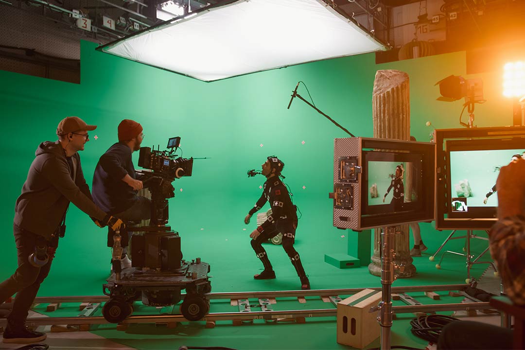Picture of a film production in a green screen studio. There is one person wearing a motion capture suit being filmed by two camera operators using a dolly. 