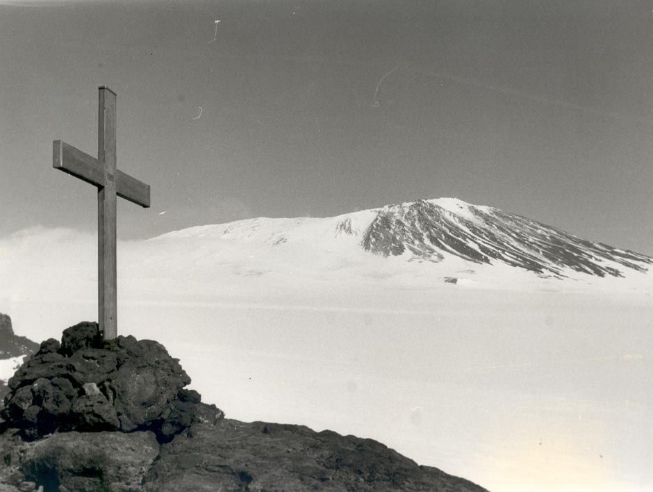 A wooden cross in the foreground. In the background is Mt Erebus covered in snow.