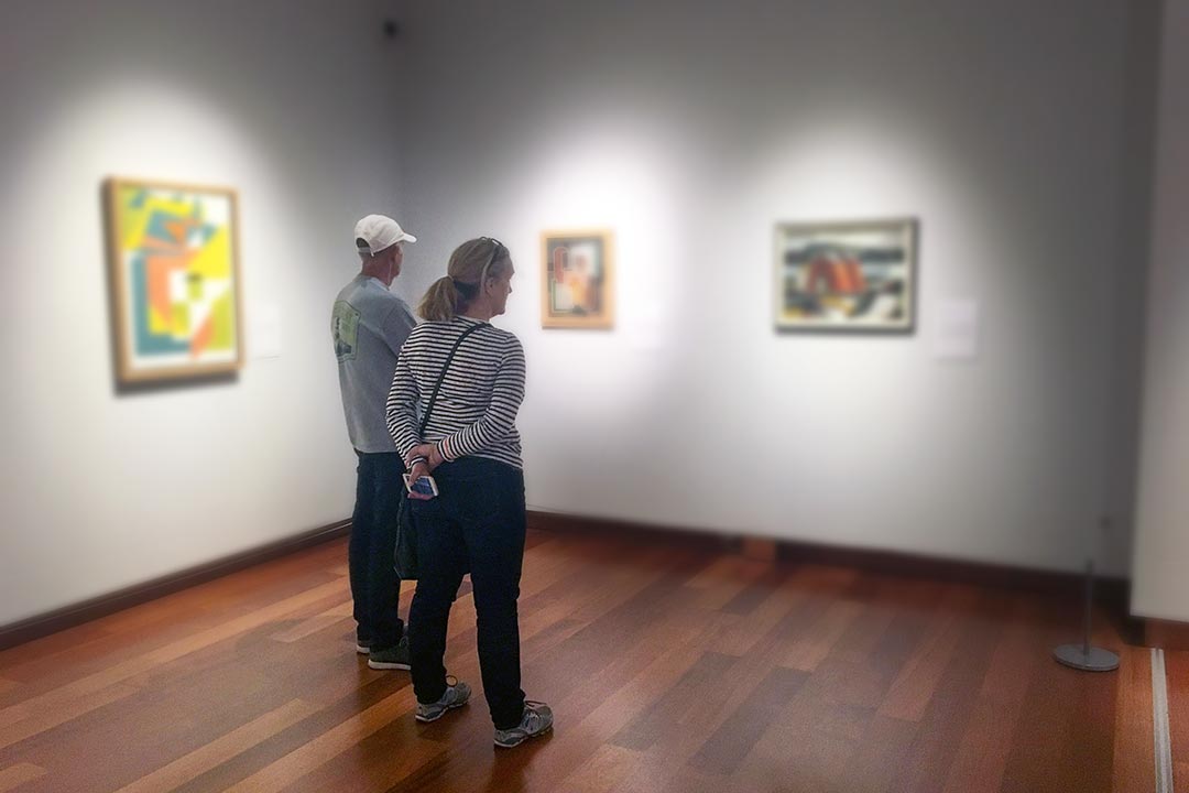 Two people looking at three paintings hung on a white gallery wall.
