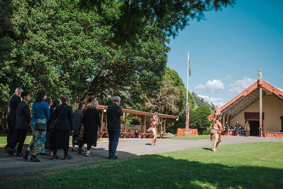 Group of people walking onto a marae. They are being met by two people performing a wero (challenge)