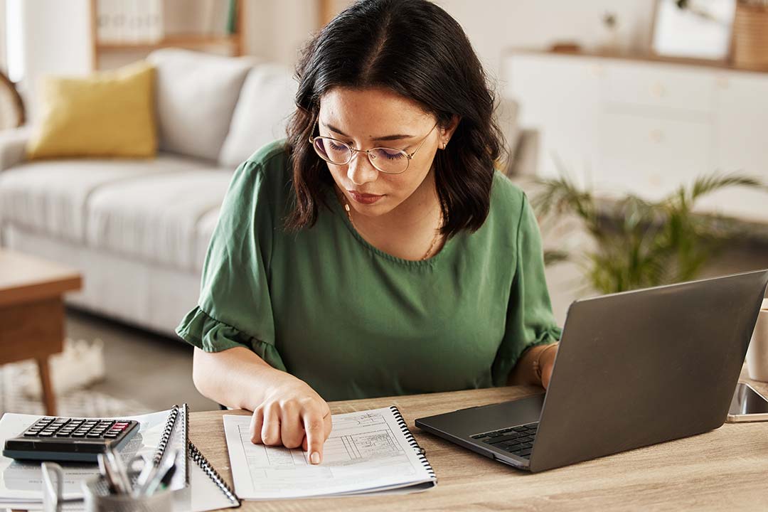 Woman in a green top looking at a piece of paper next to her laptop and calculator.