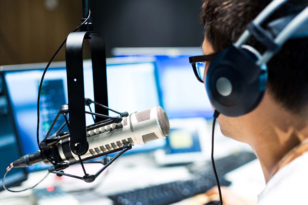 Person wearing headphones sitting in front of a radio microphone on a stand. There is a computer screen visible in background.