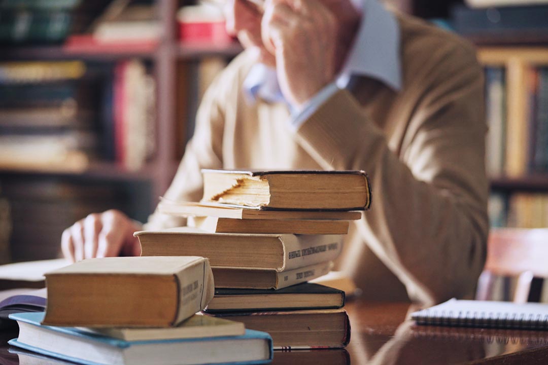 Person reading while sitting at a desk covered with stacks of books.