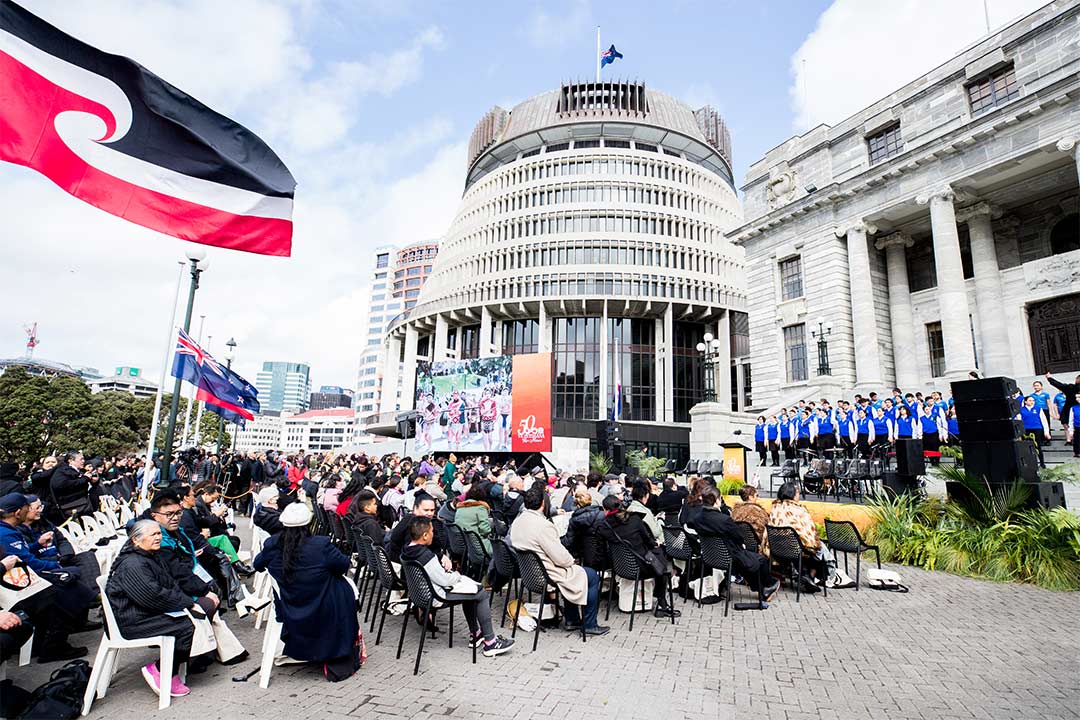 Group of people seated outside the Beehive, they are facing a stage where a group of students in blue uniforms perform for the anniversary of Te Petihana Reo Maori.