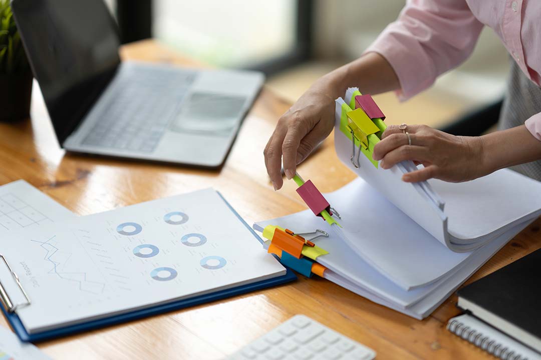 A person looks through stacked paper files. A paper on the desk shows graphs