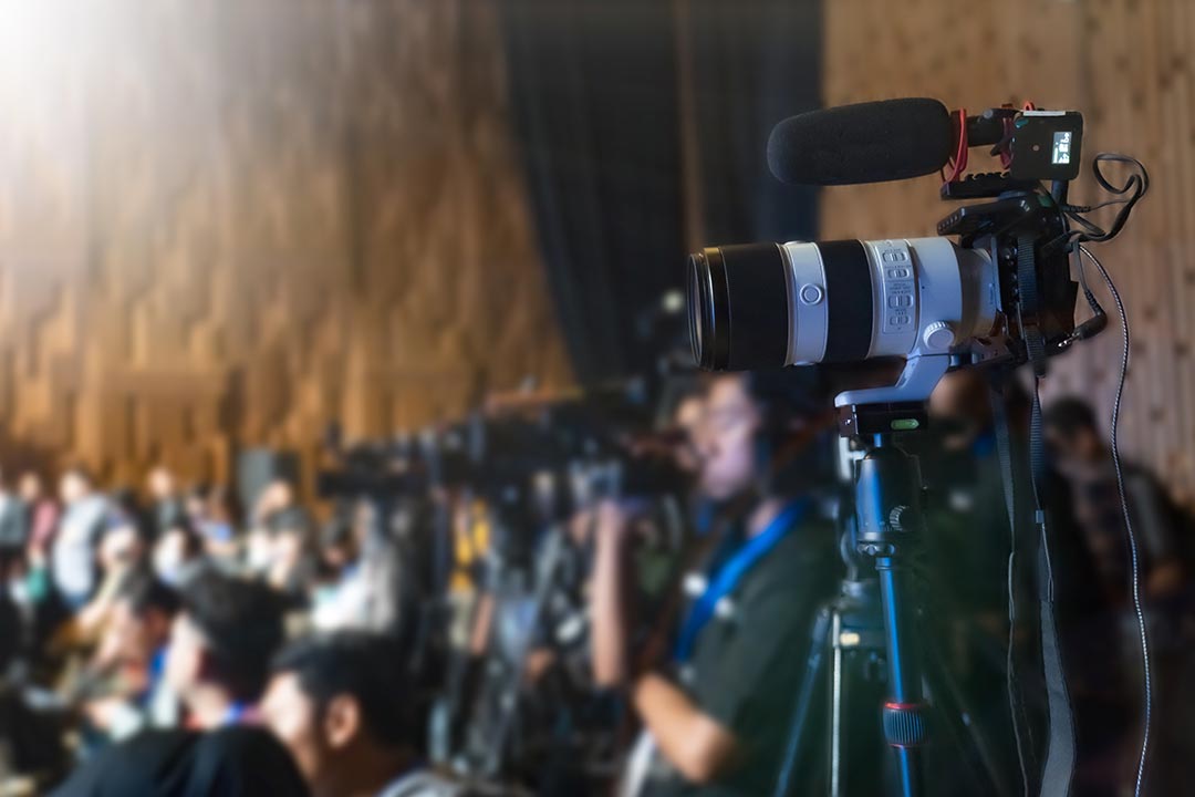 A camera on a tripod with a microphone attached in the foreground. There are people seated out of focus in the background. 