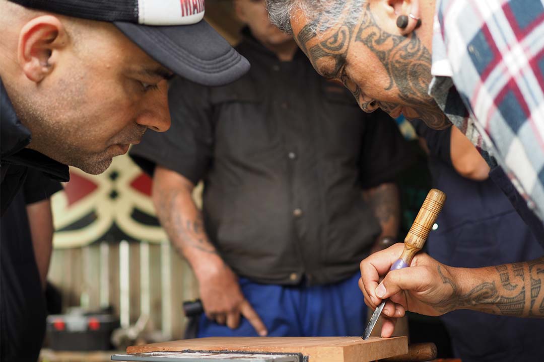 Two people leaning over a carving, one is holding a carving tool and has a facial tā moko