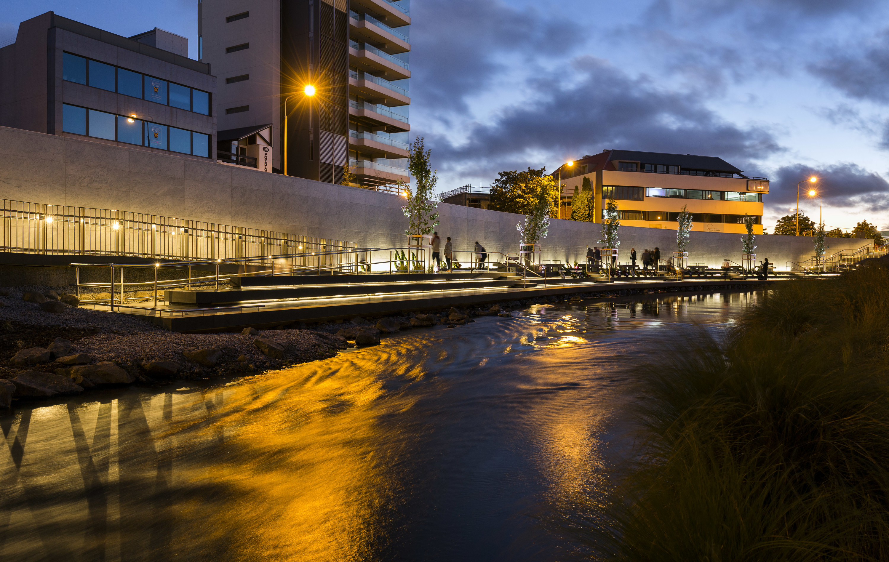 Memorial wall lit up at night with river in foreground. A few people are inspecting the wall.