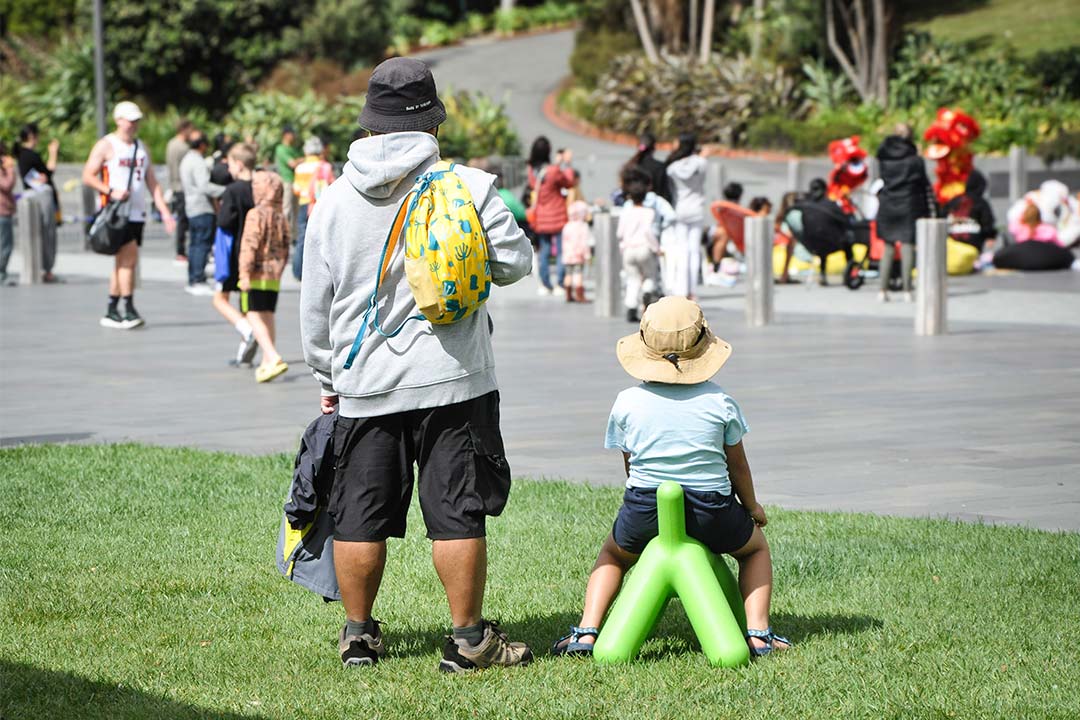 A man is standing in a park on a sunny day. His back is to the camera and he wears a yellow backpack. To the right of him is a child sitting on a green sculptural dog. They are both watching a family event taking place in the background with performers and other families.