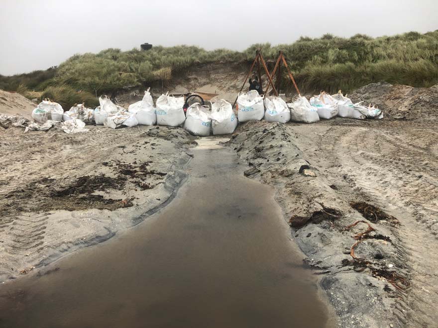 A row of white sand bags are placed along a small channel to block water running towards the excavation site from the sea.