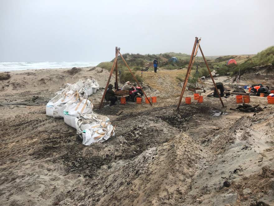 Two triangular wooden structures set up on a beach. The structures are holding another wooden box that is suspended by rope. Several orange buckets are palced around an area in the sand covered with wooden planks. There are tents and people in the background. 