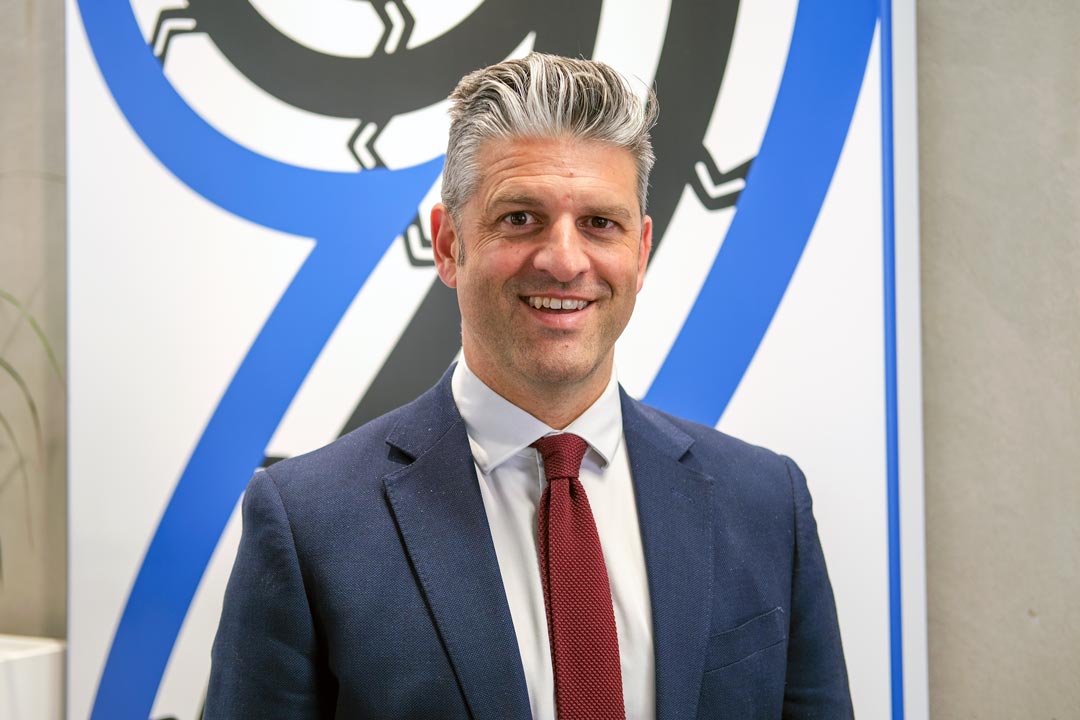Brendan smiles at the camera. He's wearing a blue suit with a red tie.