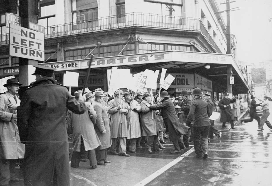 Group of men in raincoats and hats holding signs facing off against men in police uniforms