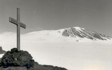 A wooden cross in the foreground. In the background is Mt Erebus covered in snow.