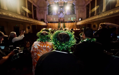 Picture of a group of people walking into the Auckland Town Hall. They are wearing 'ei katu (Cook Island wreath) made of leaves on thier head. To each side of them are members of the media holding cameras.