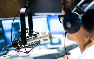Person wearing headphones sitting in front of a radio microphone on a stand. There is a computer screen visible in background.