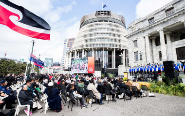 Group of people seated outside the Beehive, they are facing a stage where a group of students in blue uniforms perform for the anniversary of Te Petihana Reo Maori.