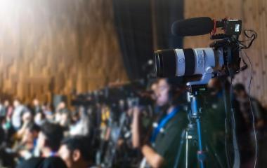A camera on a tripod with a microphone attached in the foreground. There are people seated out of focus in the background. 