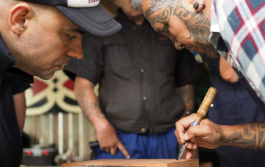 Two people leaning over a carving, one is holding a carving tool and has a facial tā moko