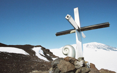 A metal cross with Mt Erebus in background covered in snow.