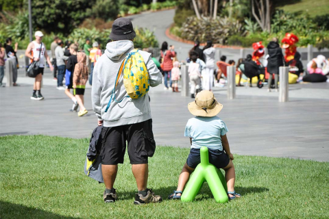 A man is standing in a park on a sunny day. His back is to the camera and he wears a yellow backpack. To the right of him is a child sitting on a green sculptural dog. They are both watching a family event taking place in the background with performers and other families.