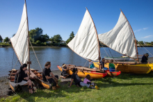People sitting around 3 waka vessels by the water