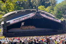 A view overlooking the Te Matatini main stage. There is a group of performers of the stage which has elements of a traditional wharenui, with a large crowd in front of them.