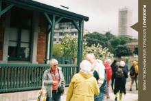 People in raincoats stand in front of a building talking in groups. A scaffolded building is in the background. The far right side has a brown banner reading 'Pukeahu National War Memorial Park'