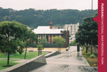 A photograph looking down a pavement to a brick building. On the left side are terraced gardens with trees. A red banner reading 'Pukeahu National War Memorial Park' is on the far right side of the tile.
