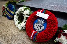 Floral wreaths rest against a black tomb. A red floral wreath is in focus, its centre holds an insignia for the Merchant Navy with the letters MN encircled by rope.